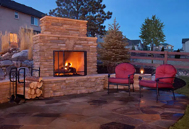 Stone outdoor fireplace on a backyard patio with seating area and lit fire creating a cozy evening atmosphere